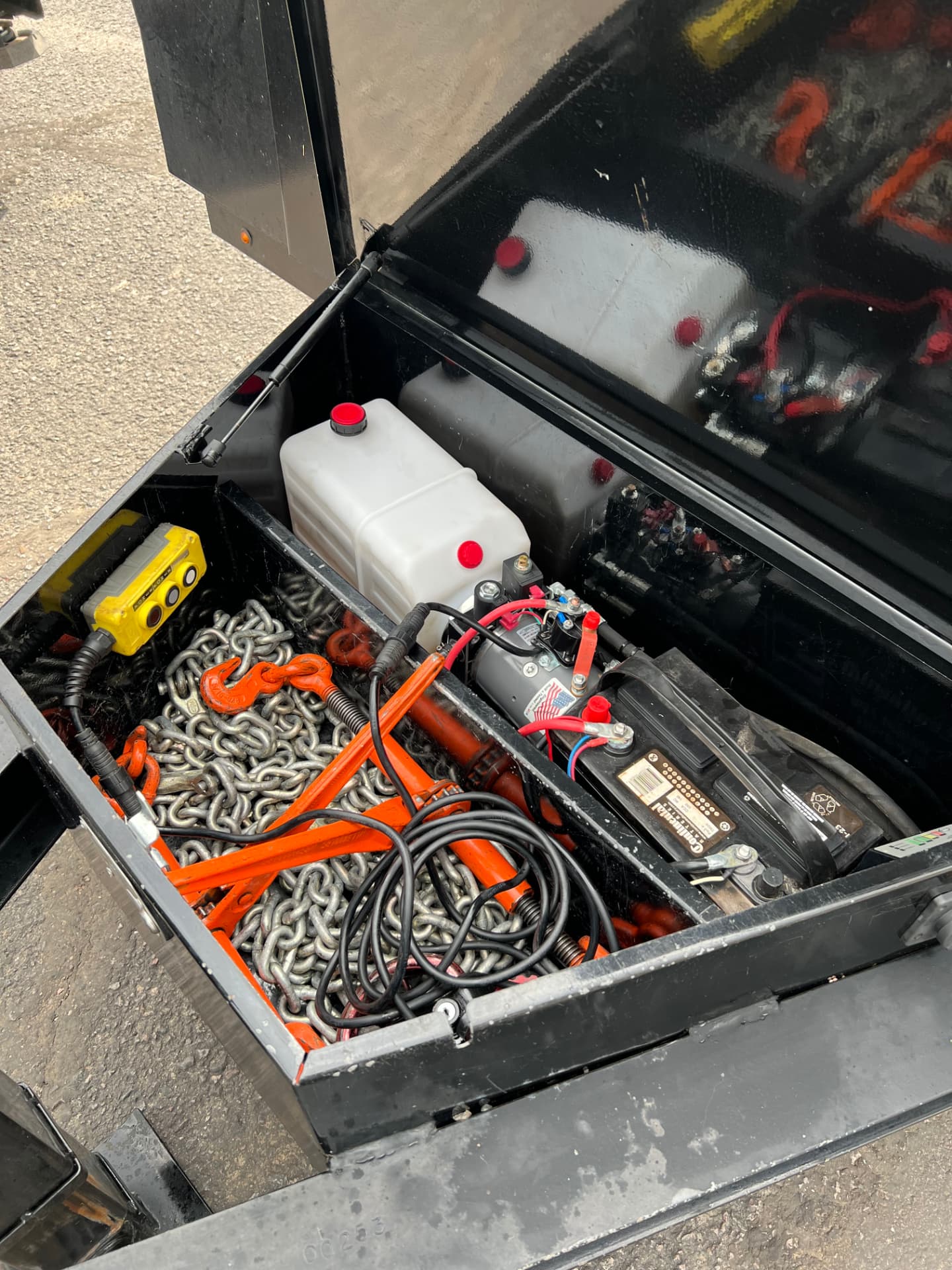 Open trailer toolbox with hydraulic pump, battery, chains and jack during a pre-trip inspection